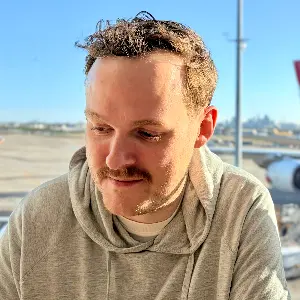Tristan, wearing a grey hoody, looking down to the left of the frame of the photo. He is bathed in sunlight from the front. Behind outside the glass is an airport runway.